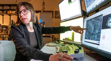 A woman wearing a black jacket gestures toward a bank of computer monitors in a lab
