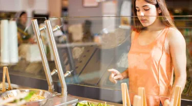 A young woman in an orange shirt chooses vegetables at a dining hall
