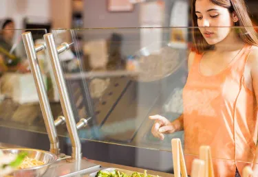 A young woman in an orange shirt chooses vegetables at a dining hall
