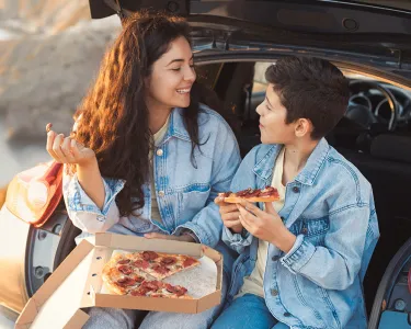 A woman and young man eat pizza sitting along the rear bumper of a car